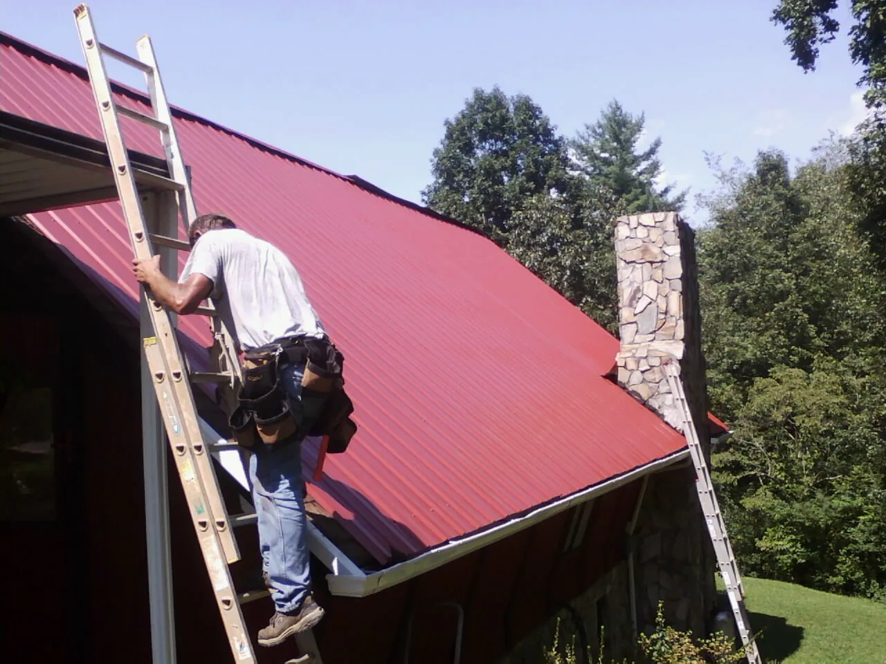 Roofer on a ladder beside a red metal roof and stone chimney — Soffit & Fascia Repair by Roofing Brothers in Rockport